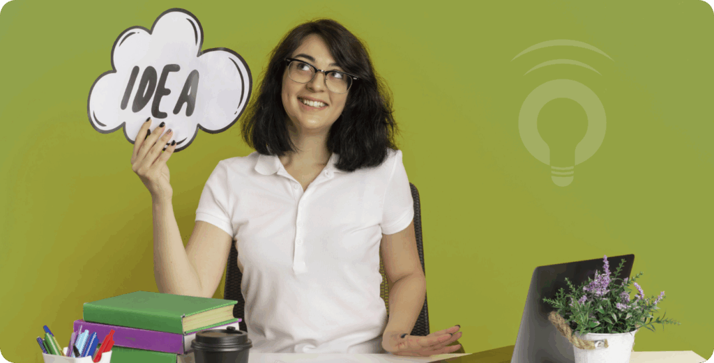 A smiling student holding a cloud-shaped sign that says "IDEA," sitting at a desk with books, a laptop, and a plant—symbolizing a classroom concept ready to be transformed into a patentable innovation using InventaIQ.