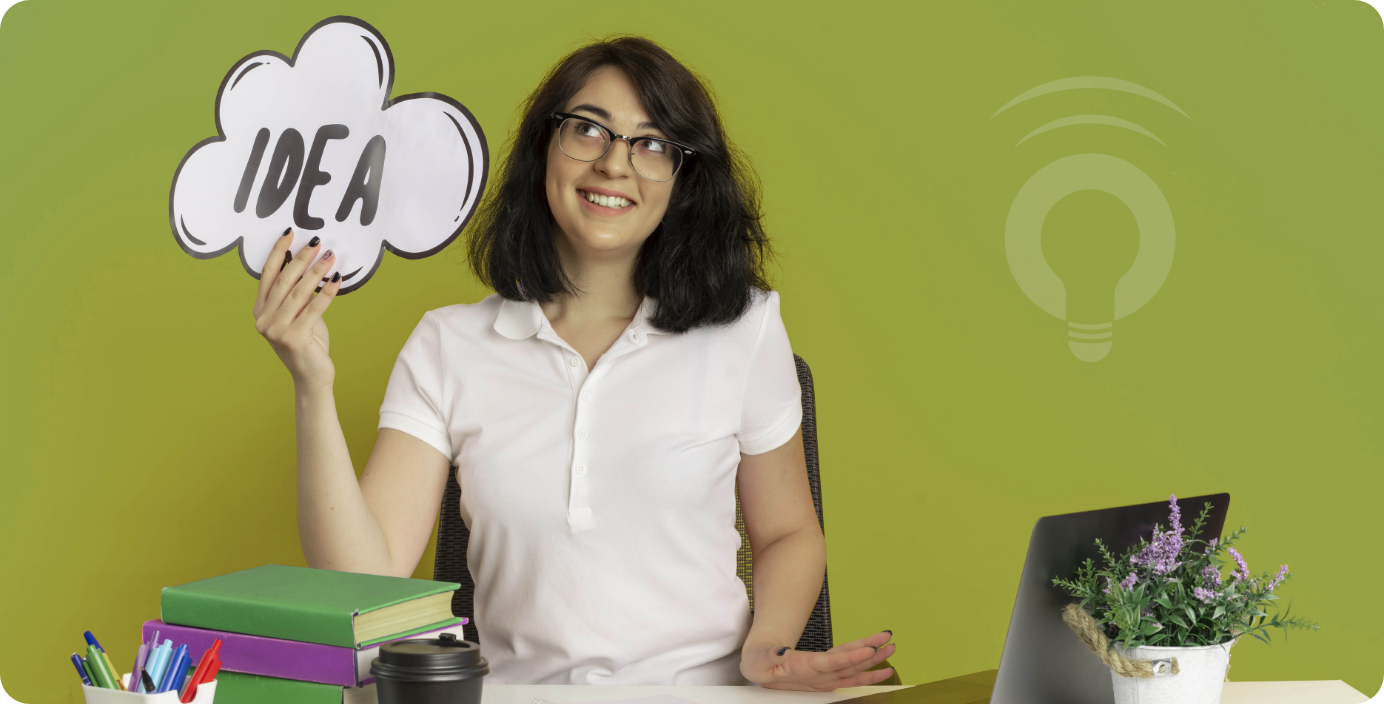 A smiling student holding a cloud-shaped sign that says "IDEA," sitting at a desk with books, a laptop, and a plant—symbolizing a classroom concept ready to be transformed into a patentable innovation using InventaIQ.